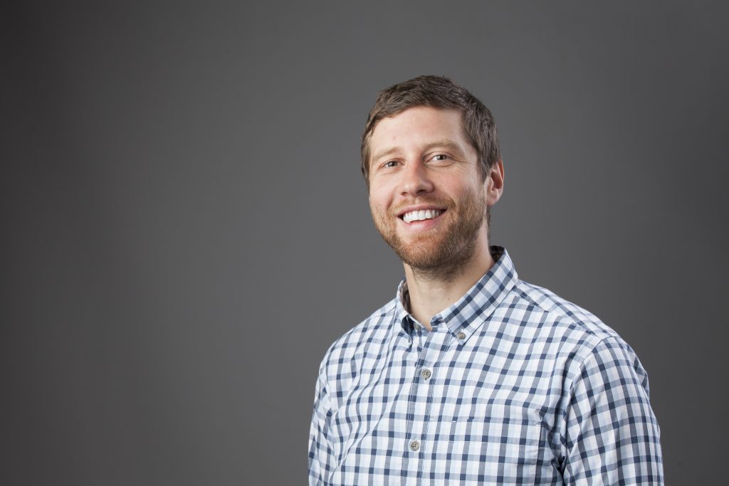 A headshot of Greg Stanfield in front of a dark grey backdrop. Greg is wearing a blue and white checked shirt.
