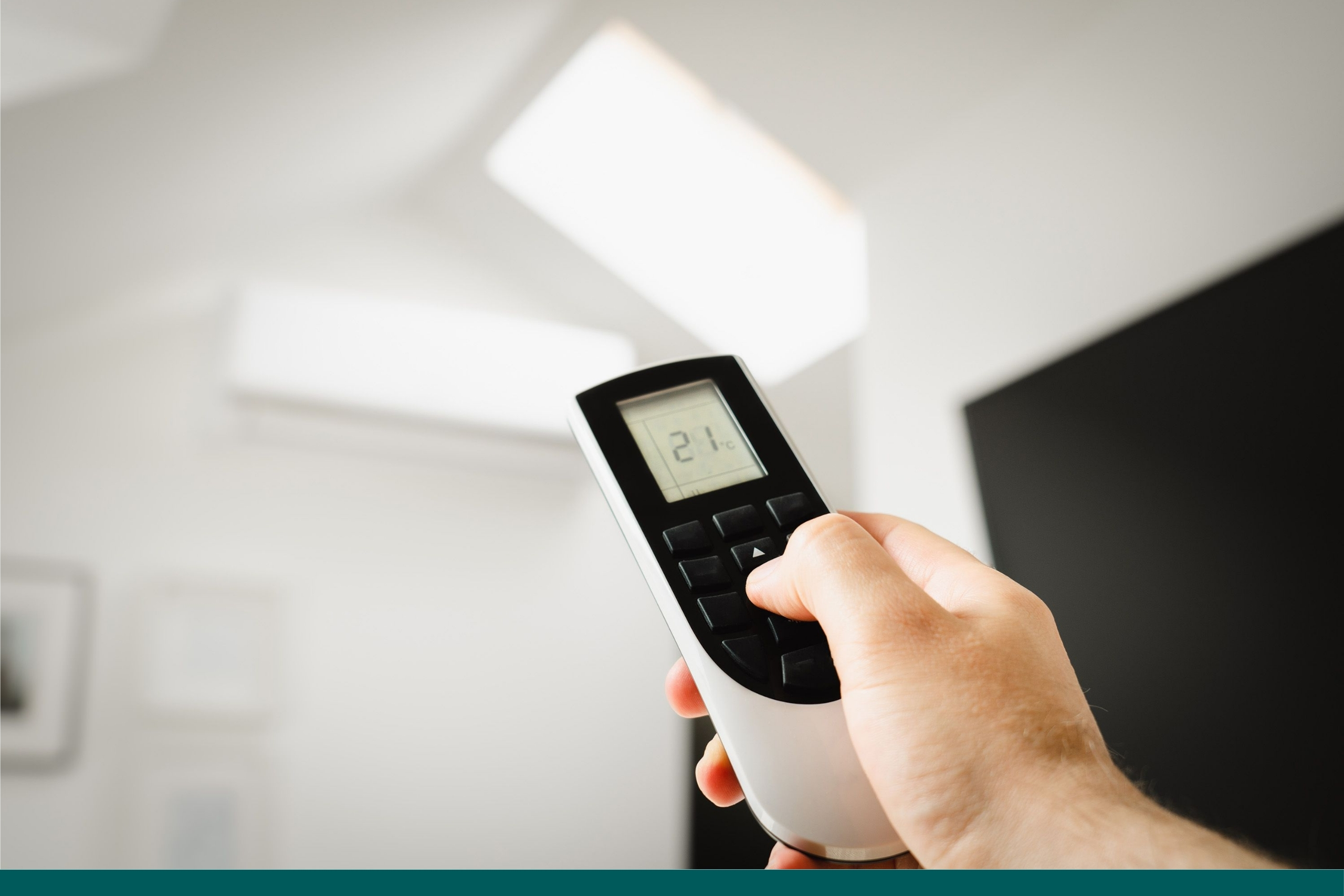 A hand holding a remote control set to 21°C, adjusting a wall-mounted air conditioning unit in a bright room